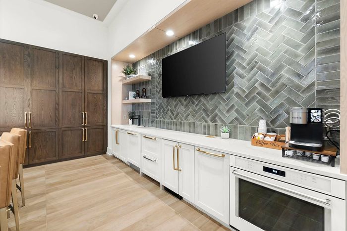 Modern kitchen area with elegant white cabinets, gold handles, and sleek appliances. Includes a chic coffee bar, stylish backsplash, and plush barstools for a cozy and inviting atmosphere.