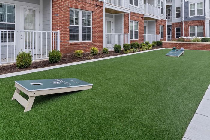Charming apartment courtyard featuring lush green lawn with cornhole game setup, surrounded by modern brick and siding architecture.