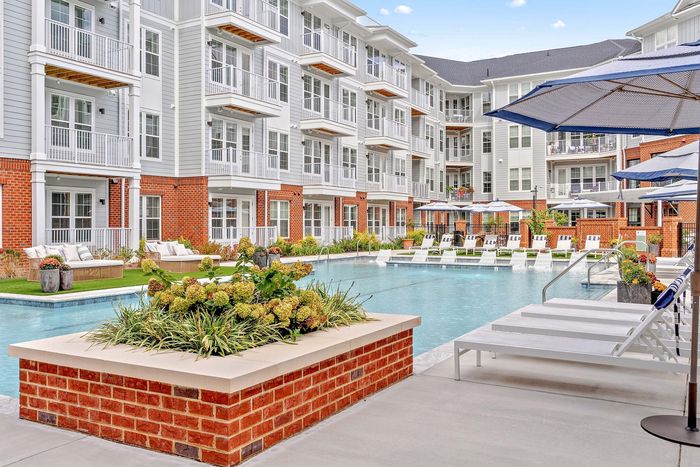 Elegant apartment complex featuring a pristine swimming pool, stylish lounge chairs, and lush landscaping, surrounded by modern multi-story buildings with private balconies.