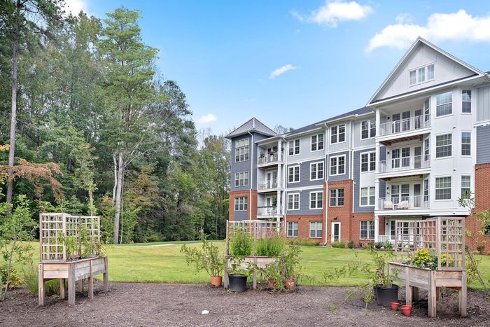 Charming brick and siding apartment complex with private balconies, surrounded by lush greenery and garden planters, under a clear blue sky.