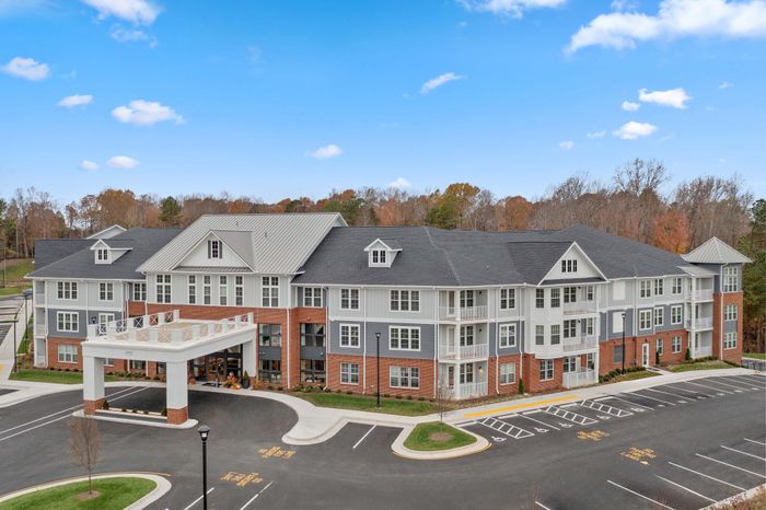 Elegant three-story apartment building with classic brick and gray siding, featuring inviting entrances and ample parking, set against a picturesque backdrop of autumn trees and a vibrant blue sky.