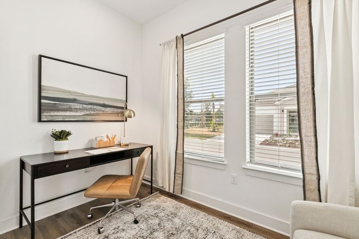 Bright home office featuring a sleek black desk, plush leather chair, and serene artwork. Large windows invite natural light, creating a peaceful and productive environment.