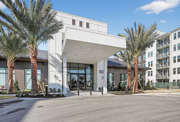 Modern apartment complex entrance with sleek architecture and lush palm trees under a clear blue sky.