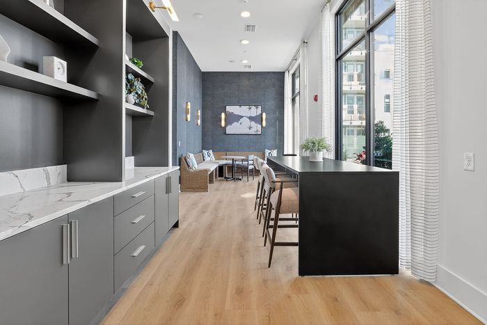 Elegant modern apartment lounge with sleek dark cabinetry, light wood flooring, and a cozy seating area by expansive windows offering abundant natural light.