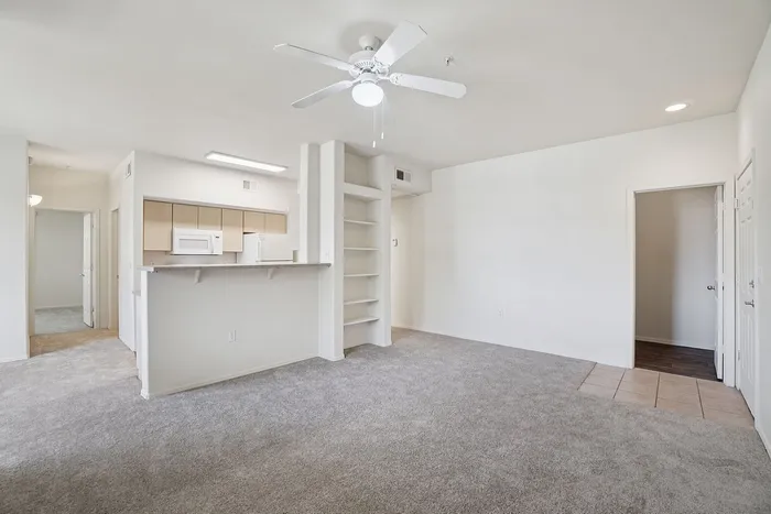 Bright, open-concept living area with plush carpet and a ceiling fan, seamlessly connected to a modern kitchen with a breakfast bar.
