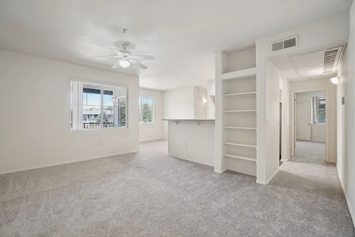 Bright and spacious living area with plush carpeting, built-in shelving, and a ceiling fan, featuring large windows for natural light.
