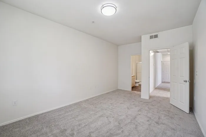 Bright and airy room with plush carpeting, modern light fixture, and hallway leading to a bathroom.