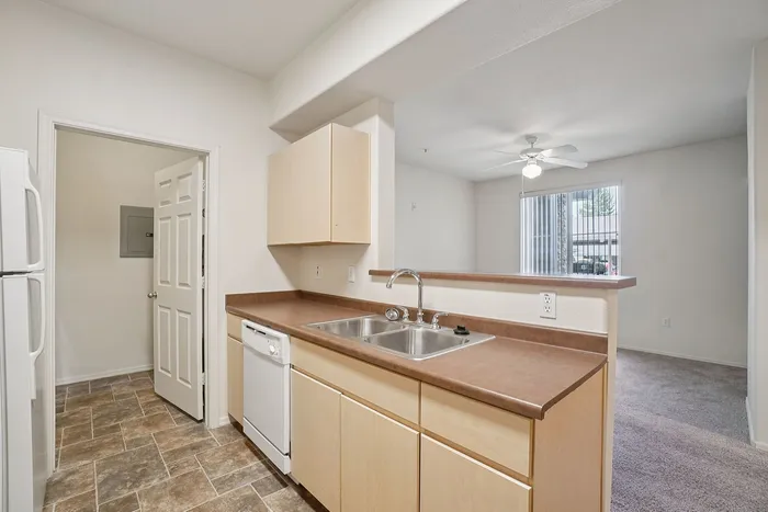Bright and inviting kitchen with sleek countertops, dual sinks, dishwasher, and open view to spacious living area featuring large window and ceiling fan.