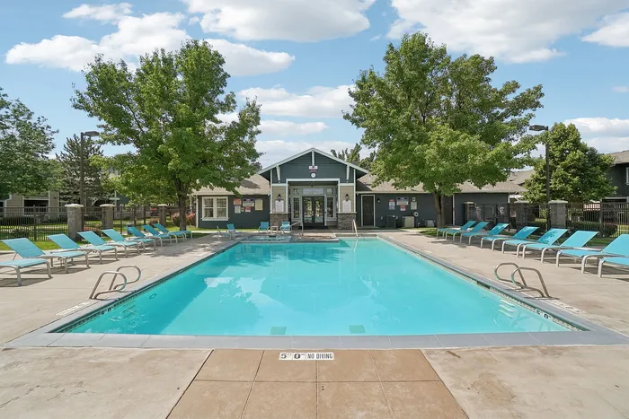 Inviting outdoor pool area with turquoise lounge chairs, surrounded by lush trees, and a charming clubhouse under a bright blue sky.