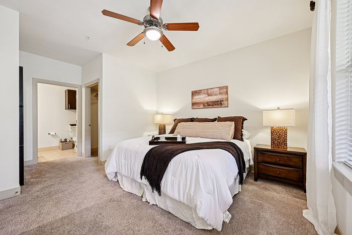 Elegant bedroom with plush carpeting, featuring a cozy king-size bed elegantly styled with luxurious bedding, beside modern wooden nightstands and a ceiling fan for ultimate relaxation.