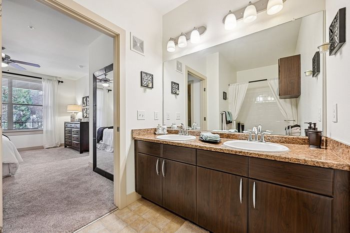 Elegant apartment bathroom with dual sinks, granite countertops, and a view into a cozy bedroom featuring soft carpeting and large windows.