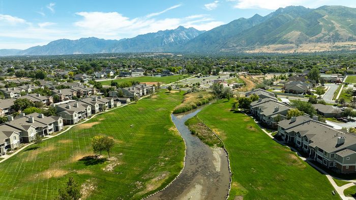Stunning aerial view of modern townhomes nestled in lush greenery, with a meandering creek and majestic mountains in the background, offering serene suburban living.