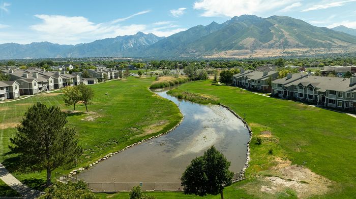 Serene aerial view of modern apartments surrounded by lush green landscape and a picturesque pond, with majestic mountains in the background offering an idyllic and peaceful setting.