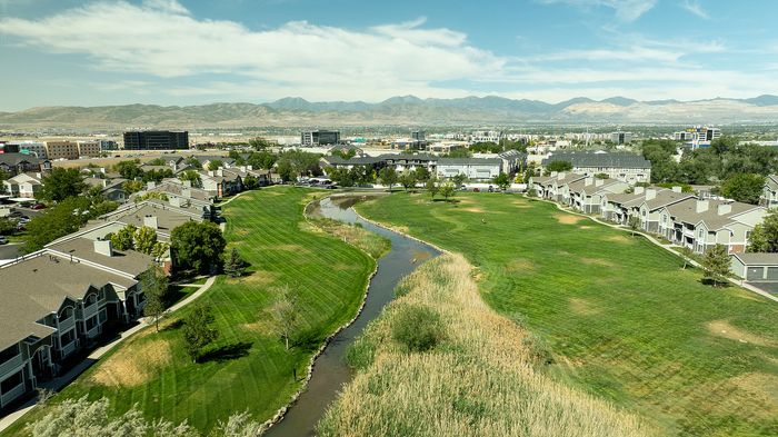 Breathtaking aerial view of a serene suburban landscape with lush green lawns, a winding stream, and modern townhouses set against a stunning mountain backdrop.
