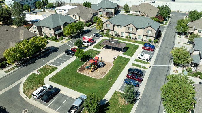 Charming aerial view of a peaceful suburban neighborhood featuring townhouses, a vibrant playground, and ample parking spaces surrounded by lush greenery.
