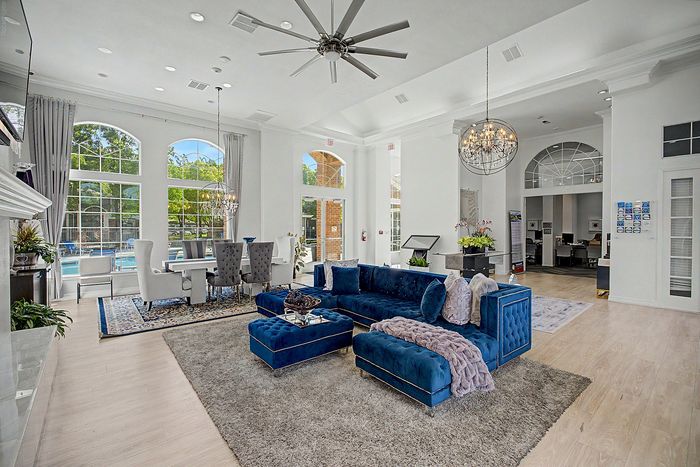 Luxurious open-concept living and dining space featuring elegant high ceilings, an opulent chandelier, plush blue velvet sectional, and expansive windows overlooking a beautiful pool area.