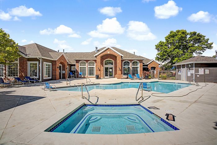 Inviting recreational area featuring a sparkling swimming pool and jacuzzi, complemented by modern lounge seating and a charming brick clubhouse under a clear blue sky.