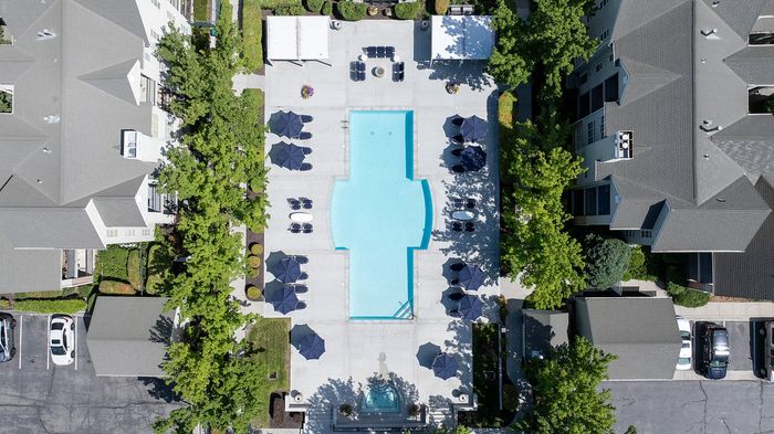 Bird's-eye view of a luxurious apartment community featuring a large, elegant swimming pool surrounded by sun loungers and shaded areas, nestled within lush greenery.