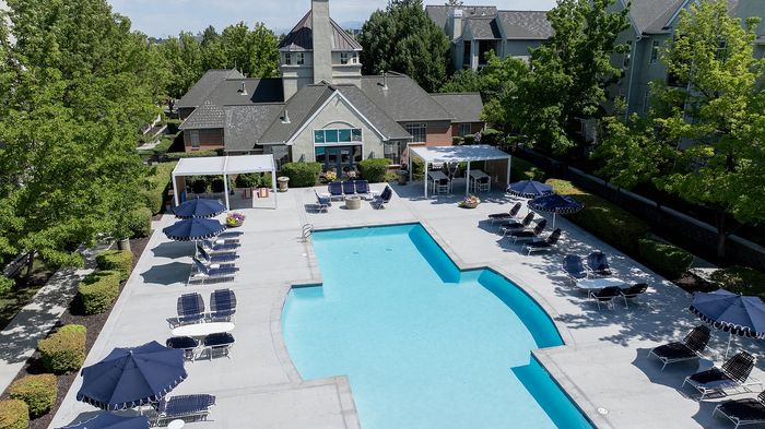 Luxurious apartment community featuring a large, inviting swimming pool with chic lounge chairs and shaded umbrellas, surrounded by lush greenery and modern architecture.