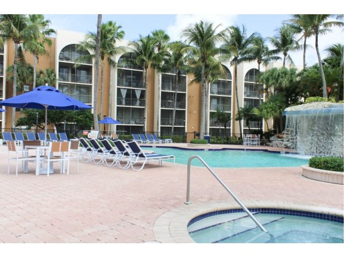 Aventura Harbor Apartments, exterior, sparkling blue pool, water fall, palm trees, umbrella covered picnic tables, clubhouse exterior