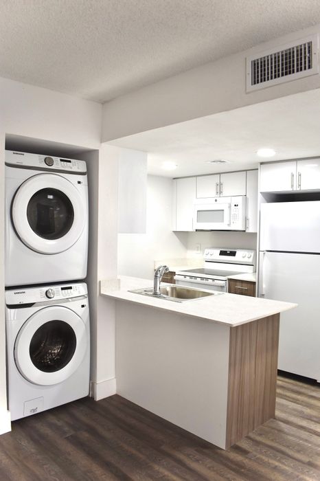 Modern kitchen with sleek appliances, stacked washer and dryer, light countertops, and wood flooring.