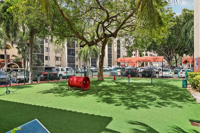 Vibrant outdoor playground with a lush green lawn, surrounded by trees and modern apartment buildings, offering a serene escape for families.