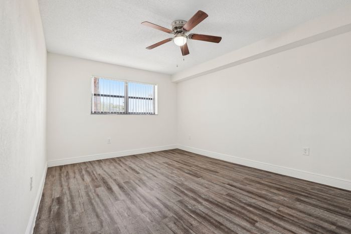 Bright and spacious bedroom featuring elegant wood flooring and a modern ceiling fan, complemented by ample natural light through a large window with blinds.