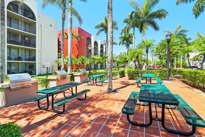 Vibrant apartment complex with lush palm trees, outdoor picnic area, BBQ grills, and sunny courtyard.