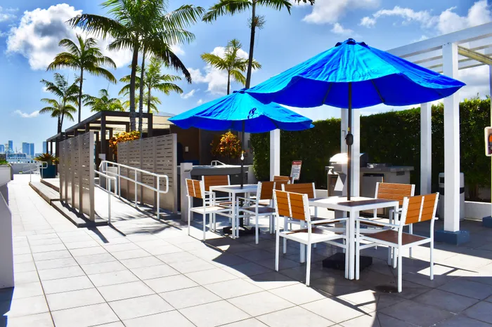 Chic outdoor terrace with bright blue umbrellas, modern seating, and lush palm trees under a sunny sky, perfect for relaxation.