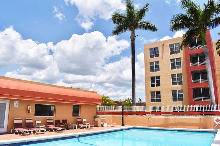 Vibrant apartment complex with a spacious outdoor pool area, sun loungers, and towering palm trees under a bright blue sky.