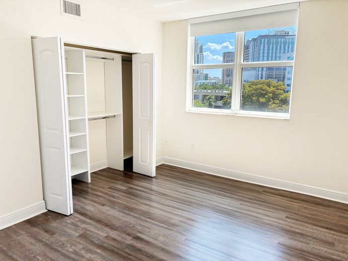 Bright, spacious bedroom with sleek wooden flooring, large window with city view, and a built-in closet with shelves and hanging space.
