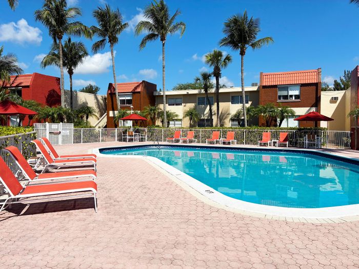 Inviting swimming pool area surrounded by palm trees and modern apartment buildings with vibrant red sun loungers and umbrellas, perfect for relaxation.