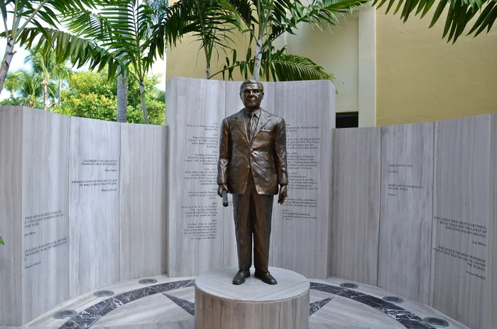 Elegant bronze statue of a suited man surrounded by tall palm trees and engraved stone walls under a clear sky, evoking a sense of dignity and history.