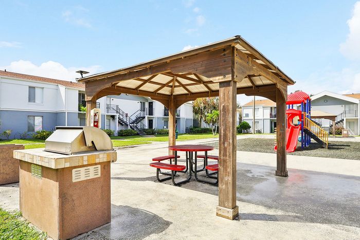 Bright community space featuring a wooden gazebo with picnic seating and a modern grill, surrounded by lush greenery and a vibrant playground in the background.