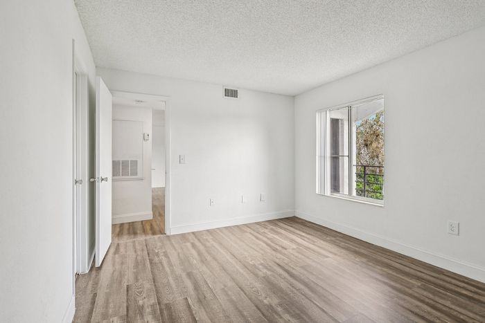 Bright and airy bedroom with sleek hardwood floors, large window offering natural light, and a white minimalist interior design.
