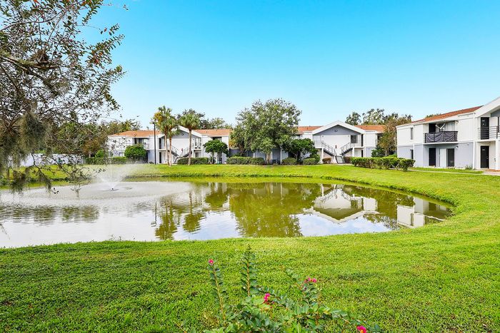 Charming apartment complex with serene pond views, lush greenery, and modern white buildings under a clear blue sky.
