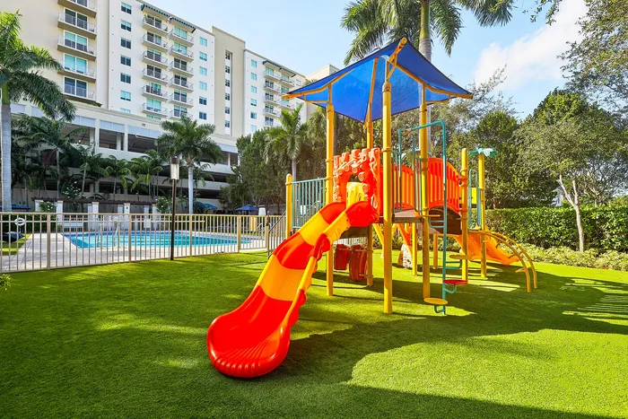 Vibrant playground with a colorful slide next to a luxurious apartment complex with a pool, surrounded by lush palm trees.