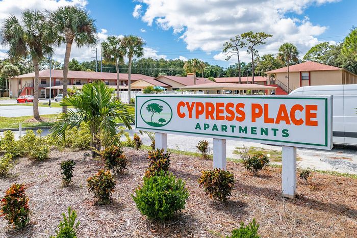 Cypress Place Apartments entrance sign surrounded by lush greenery and tropical palm trees under a vibrant blue sky.