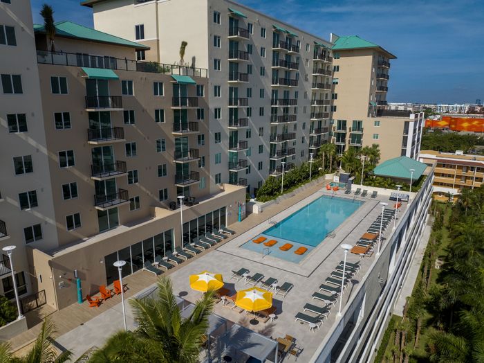 Modern apartment complex with expansive pool, sun loungers, and vibrant umbrellas surrounded by lush greenery and cityscape views.