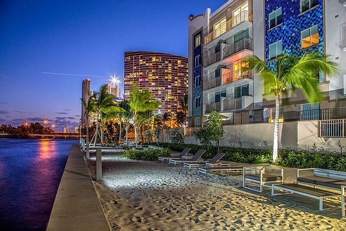 Luxurious waterfront apartment complex with modern architecture, palm trees, and lounge chairs on a sandy beach, beautifully illuminated at dusk.