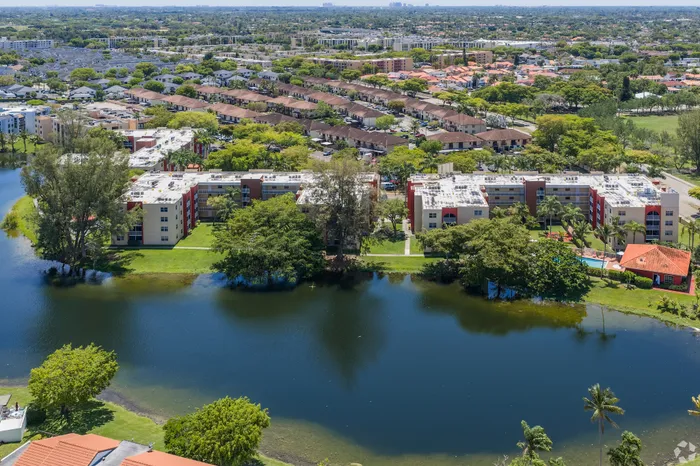 Scenic aerial view of a vibrant apartment complex nestled by a serene lake, surrounded by lush greenery and a sprawling residential neighborhood.