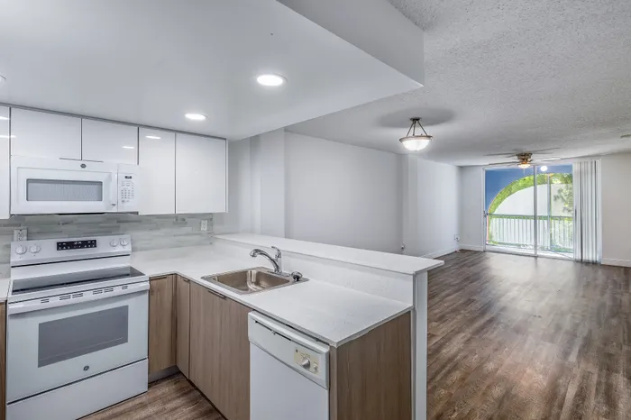 Bright and modern kitchen with sleek white cabinets and appliances opens into a spacious living area with natural light from large balcony doors.
