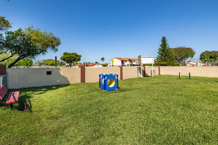 Inviting green grassy area with a playful blue and yellow structure, bordered by a privacy wall and shaded trees, offering a tranquil outdoor space in a serene neighborhood.
