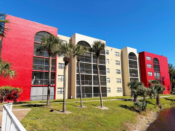 Vibrant apartment complex with striking red and beige facades, surrounded by lush palm trees and clear blue skies.