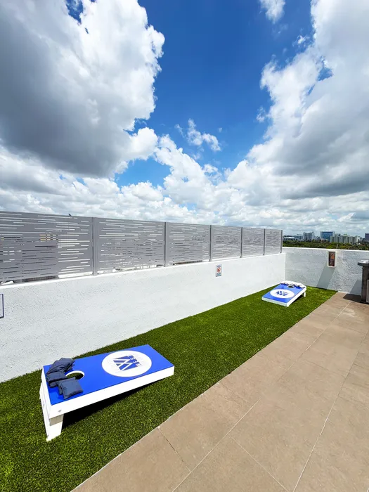 Inviting rooftop space with a cornhole game on lush artificial grass, set against a backdrop of a vibrant blue sky with fluffy clouds.