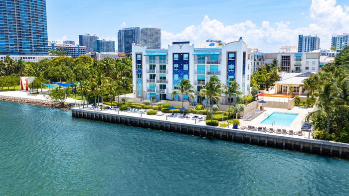 Luxurious waterfront apartment complex with vibrant blue accents, lush tropical landscaping, and a sparkling pool overlooking the serene waterway.