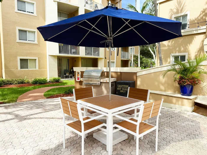 Inviting outdoor patio with modern furniture and grill, set under a large blue umbrella, surrounded by lush greenery and charming apartments.