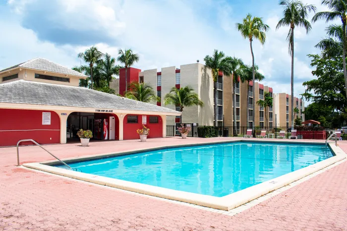 Inviting apartment complex with a sparkling outdoor pool, surrounded by lush palm trees and modern buildings, offering a tropical oasis ambiance.