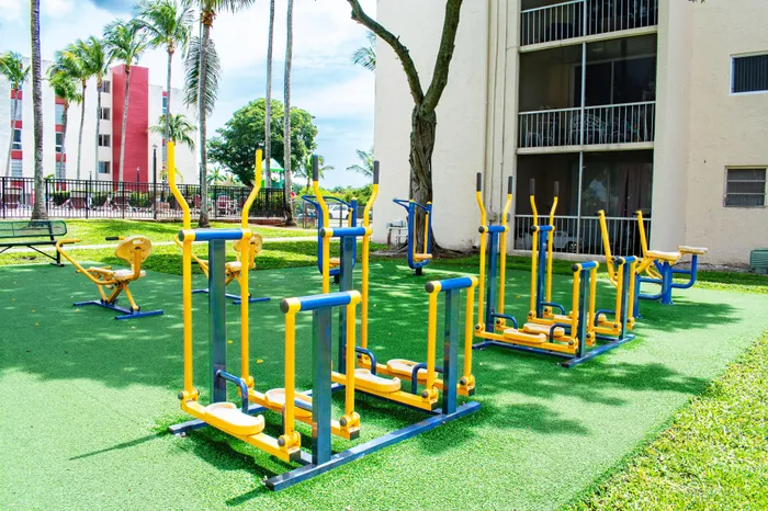Outdoor fitness area with vibrant yellow and blue exercise equipment surrounded by lush greenery and a residential building backdrop.