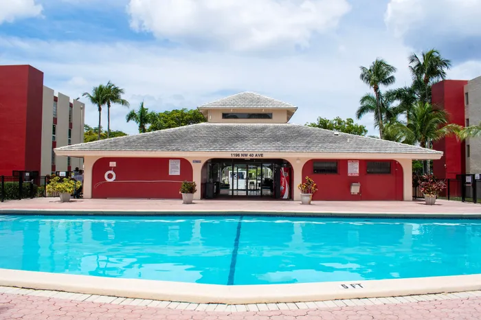Inviting apartment complex with a sparkling outdoor pool, surrounded by lush palm trees and vibrant red buildings, perfect for relaxation and leisure.